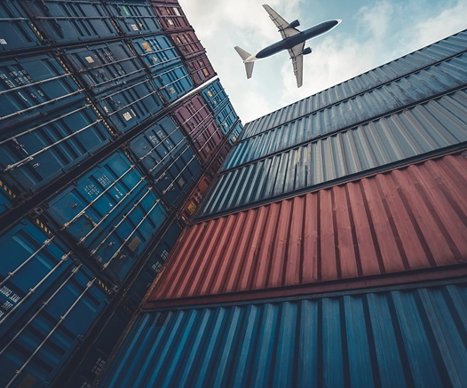 Widok od dołu na ułożone w stosy niebieskie i czerwone kontenery transportowe, nad którymi przelatuje samolot na tle zachmurzonego nieba, oddający ruch i skalę.