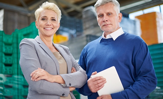 Dwie osoby stoją pewnie w magazynie. Kobieta ma na sobie szarą marynarkę, uśmiecha się i trzyma ręce skrzyżowane. Mężczyzna trzyma tablet i ma na sobie niebieski sweter.