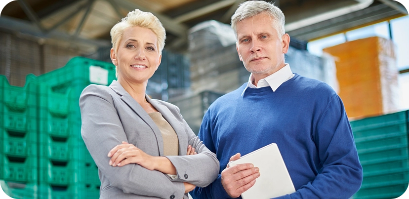 Dwoje pewnych siebie profesjonalistów stoi w magazynie. Kobieta w szarym garniturze krzyżuje ręce, a mężczyzna w niebieskim swetrze trzyma tablet.